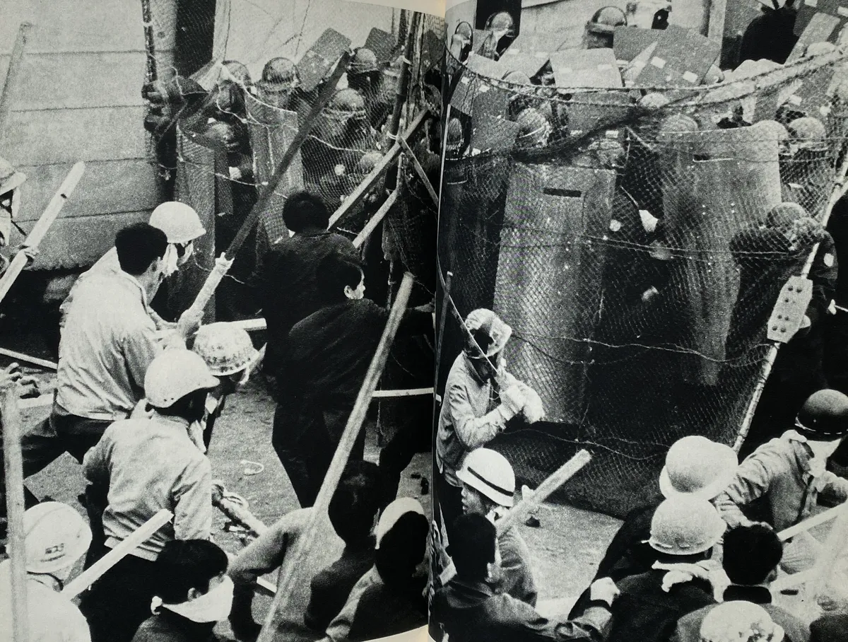 Fotografia en blanc i negre. Manifestants atacant a la policia. Porten tots uns característics cascos d'obra amb caràcters japonesos escrits a sobre. Ataquen amb pals gruixuts, mentre la policia es defensa amb escuts que semblen d'acer o ferro.