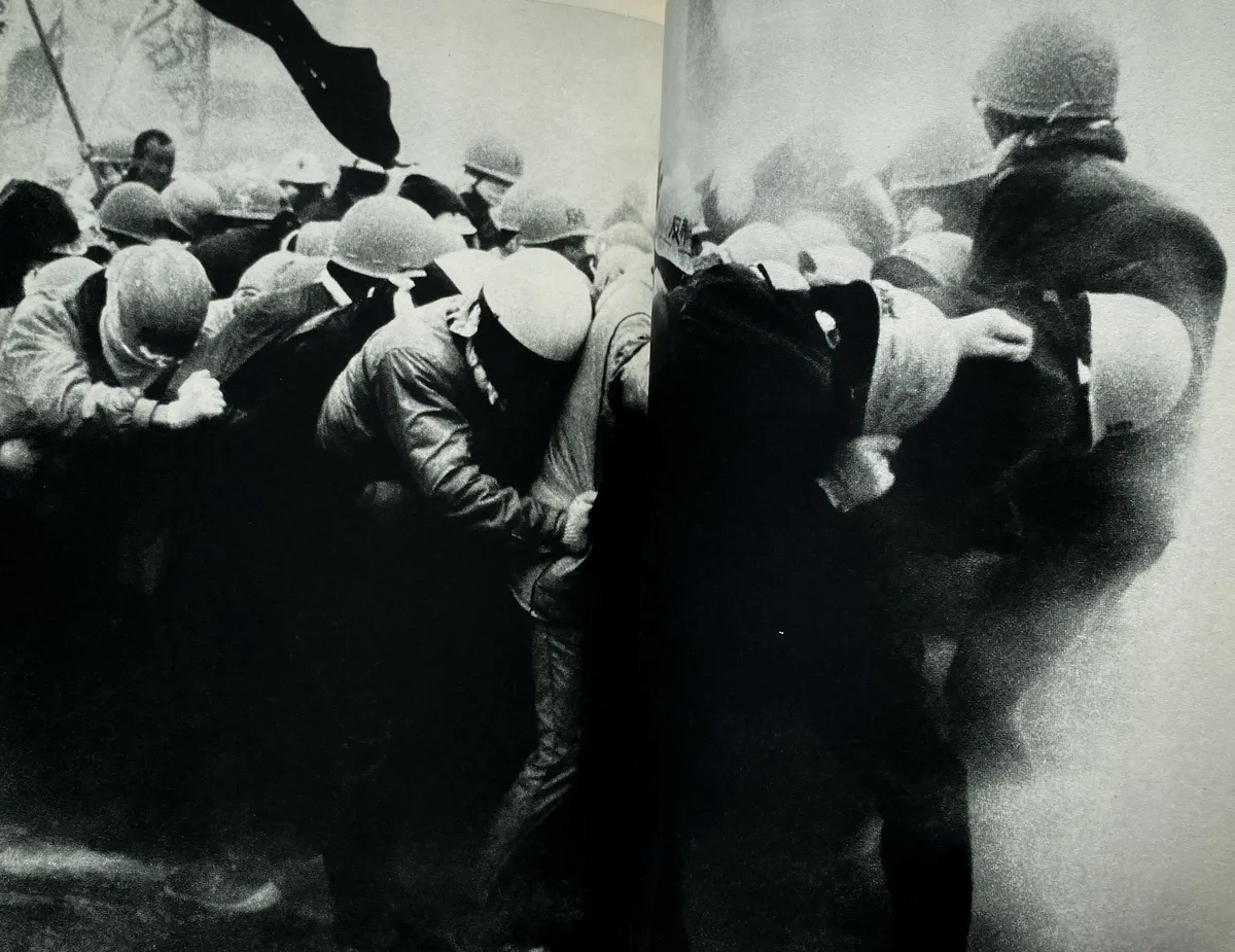 Fotografia en blanc i negre. Manifestants s'agafen entre ells mentre un raig d'aigua els cau a sobre. Tots porten els característics cascos d'obra amb caràcters japonesos.