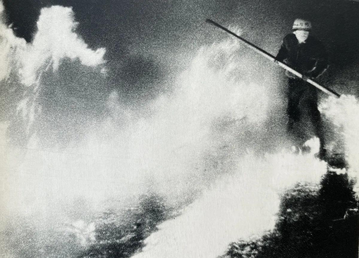 Fotografia en blanc i negre. Un manifestant, amb el seu casc blanc i un pal gran a les mans, travessa una barricada que crema produint unes flames molt més grans que ell.